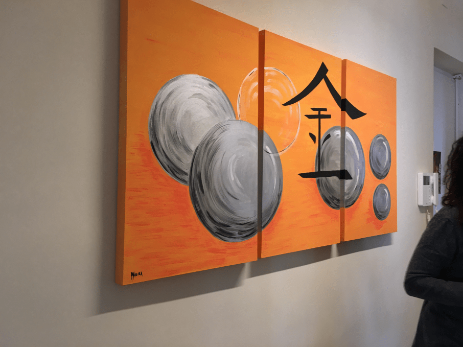 Quadro asiatico tripartito con sfondo arancione, un grande ideogramma cinese al centro e delle sfere grigie attorno