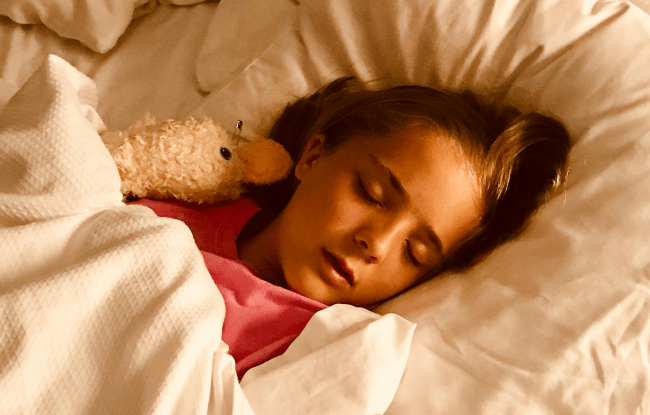 Una bambina dorme tranquillamente su un letto con lenzuola bianche, abbracciando un peluche a forma di papera. Indossa una maglietta rosa e ha i capelli biondi sciolti sul cuscino.