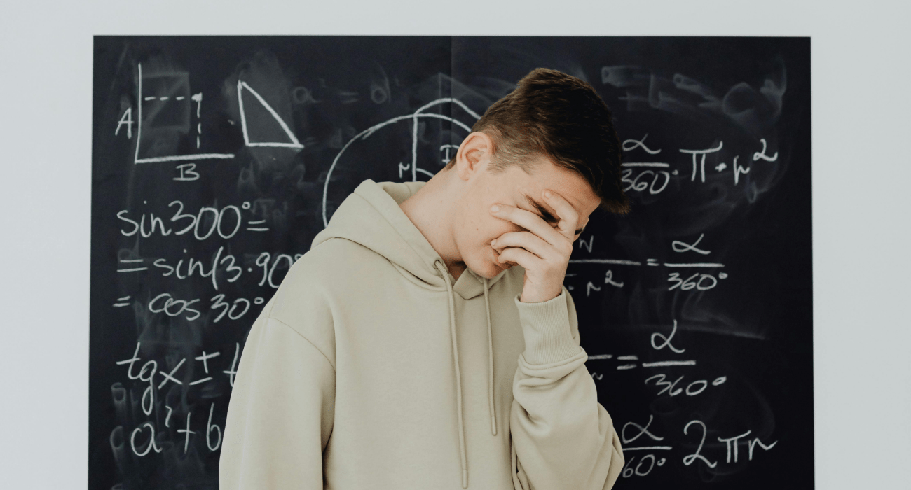 Un ragazzo in felpa davanti ad una lavagna nera rimane deluso della sua prestazione scolastica e tiene una mano sul volto per coprirlo. Ferite karmiche e rinascita.