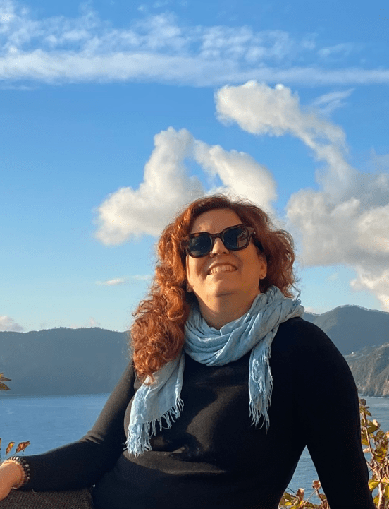 Raffaela Marescalco sorridente, con occhiali da sole dalla montatura color marrone scuro, posa davanti ad un lago durante una bella giornata di sole.