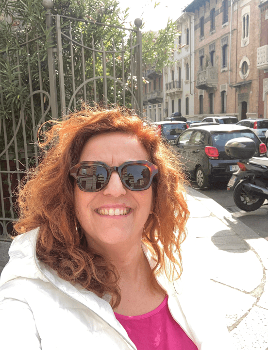 Raffaela Marescalco sorridente, con occhiali da sole dalla montatura color marrone scuro, posa davanti ad un cancello al cui interno troviamo una pianta.