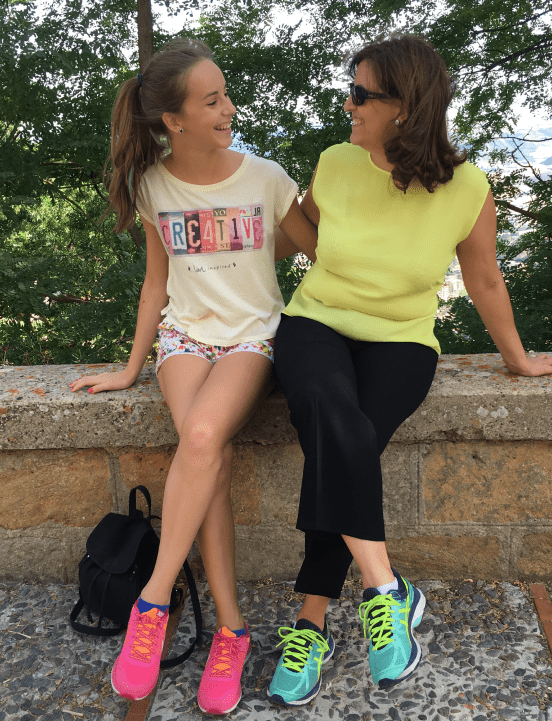 Raffaela Marescalco e sua figlia maggiore Sarah sedute su un muretto all'aperto, sorridono guardandosi negli occhi. Entrambe indossano abiti casual e scarpe da ginnastica colorate.